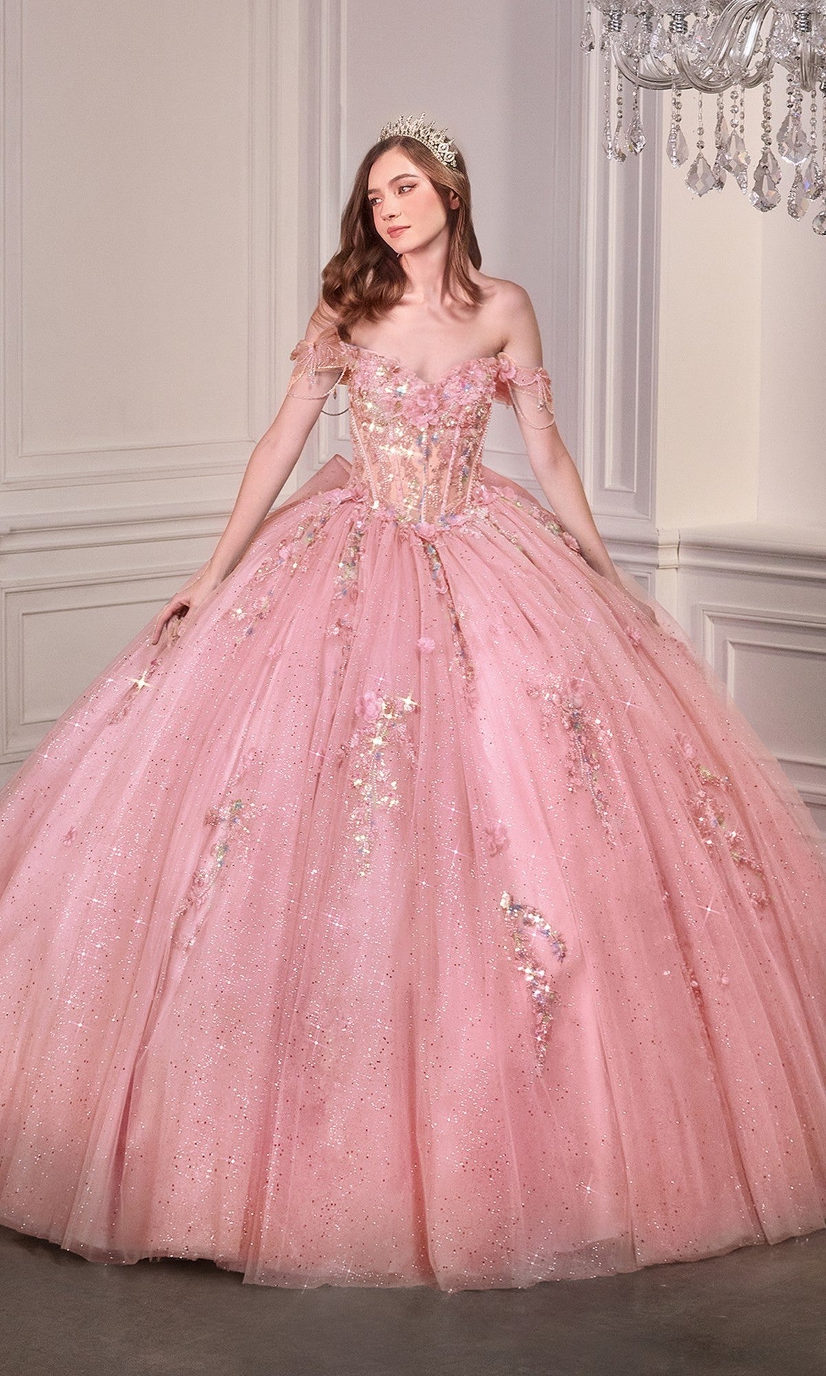 A woman in the Quinceanera Dress 15728 By Ladivine, a sparkling pink ball gown with off-the-shoulder sleeves and a tiara, stands in an elegant white-paneled room.